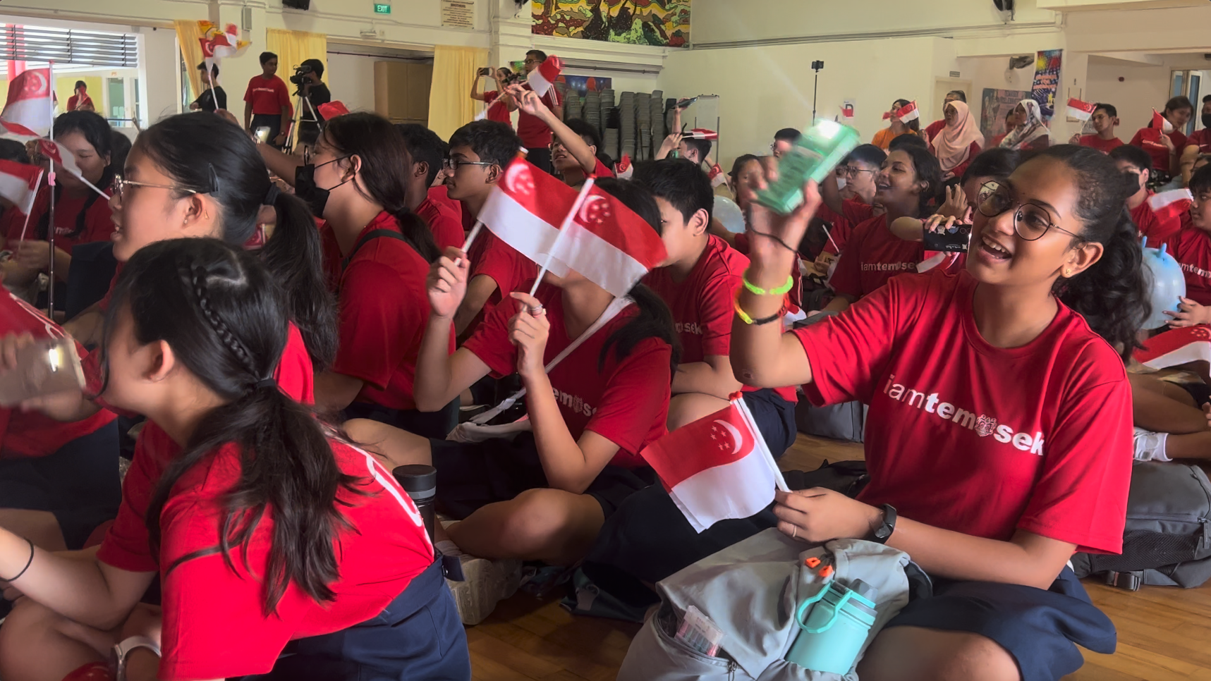 Students in red shirts wave Singapore flags while seated on a hall floor during a school celebration event.